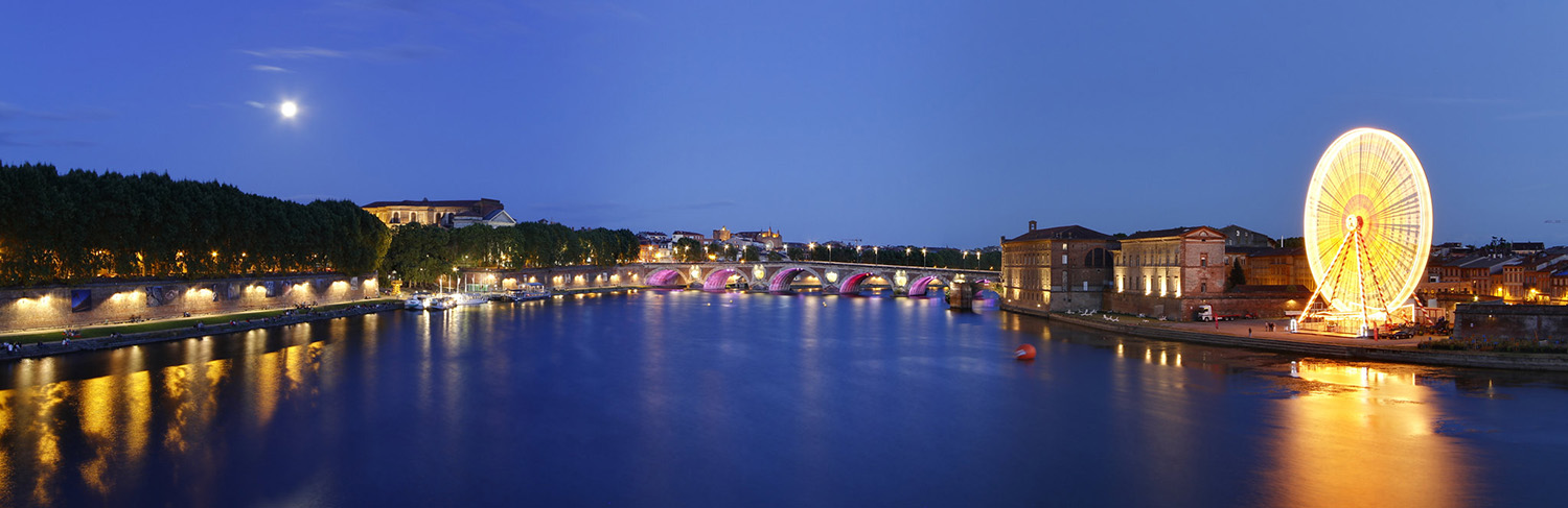 Photograph of Toulouse at night. The canal is deep blue, and the arches of the Napoleonic bridge are lit up. 